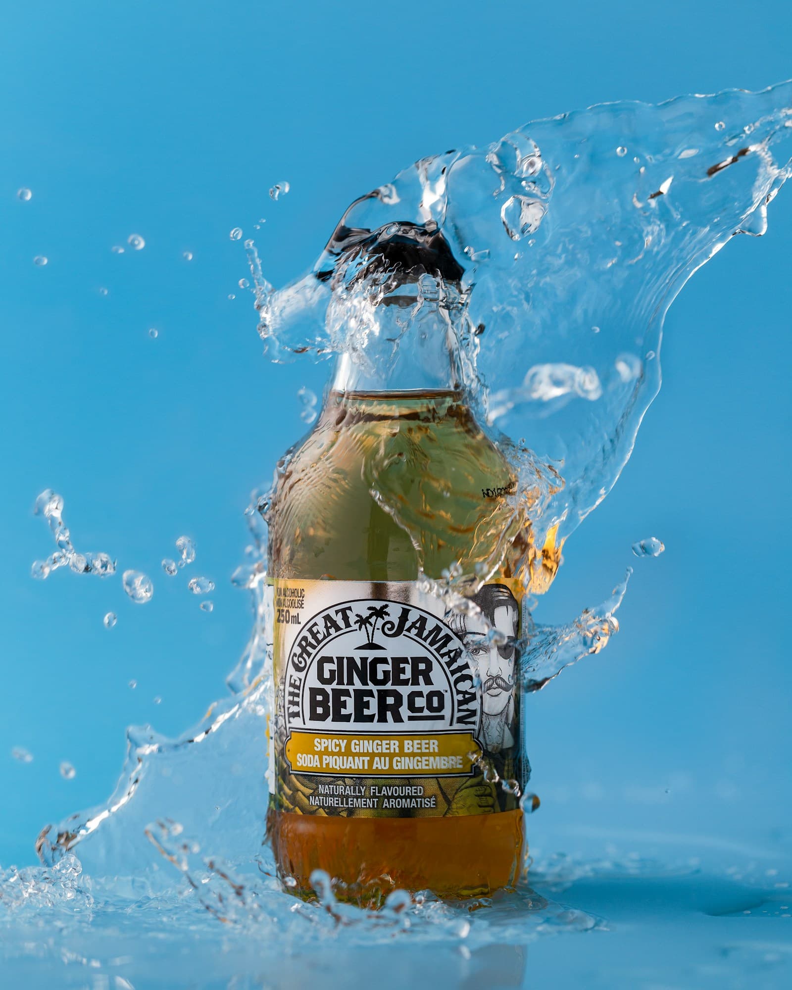 A glass bottle of ginger beer standing against a blue background is surrounded by splashes of water.